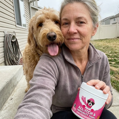 A happy customer holding a jar of ALL-ITCH & IMMUNITY Treats next to her fluffy Goldendoodle dog.